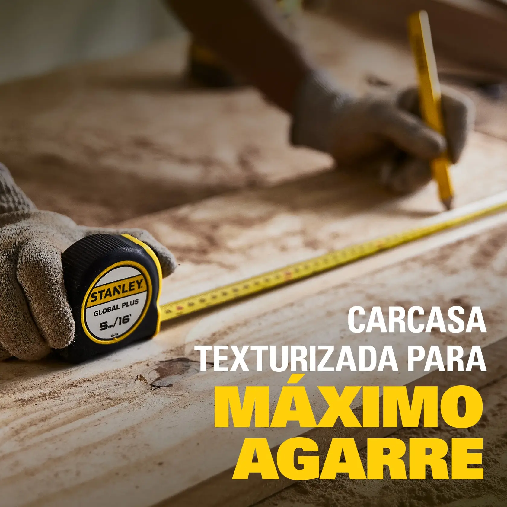 Una persona con guantes de trabajo utiliza una cinta métrica Stanley Global Plus de 5 m/16 pies sobre una superficie de madera, marcando una medida con un lápiz. El texto resalta la carcasa texturizada para máximo agarre.