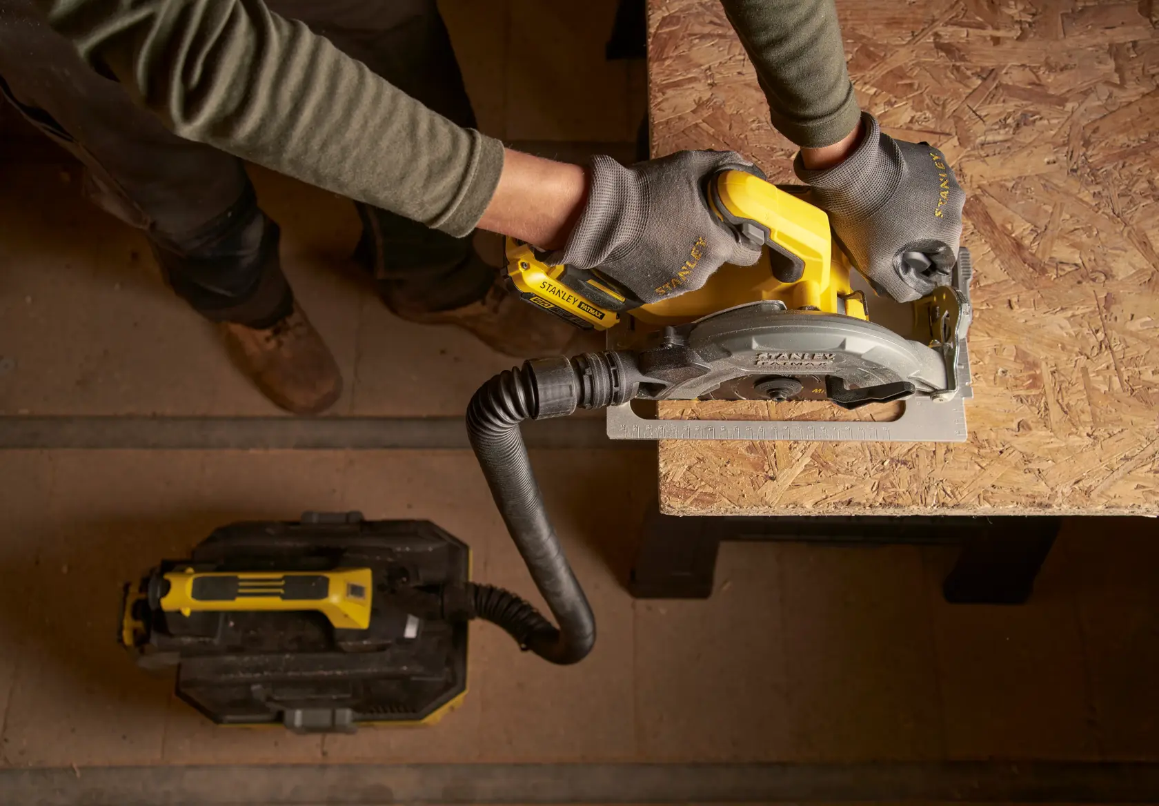 STANLEY SBC550D2K-B3 sierra circular inalámbrica en uso sobre panel de madera, conectada a extractor de polvo, vista superior.