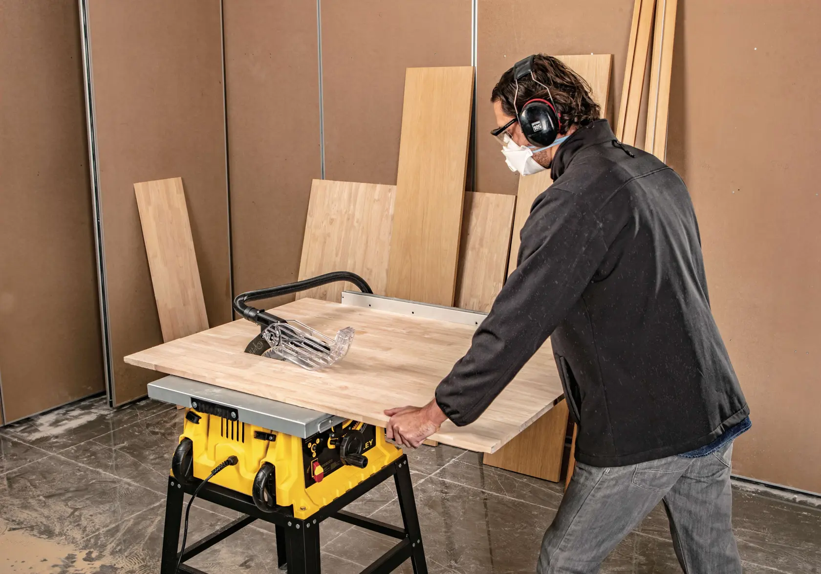 Sierra de mesa STANLEY SST1801-B3 en uso cortando un panel de madera, persona con protección auditiva, taller interior.