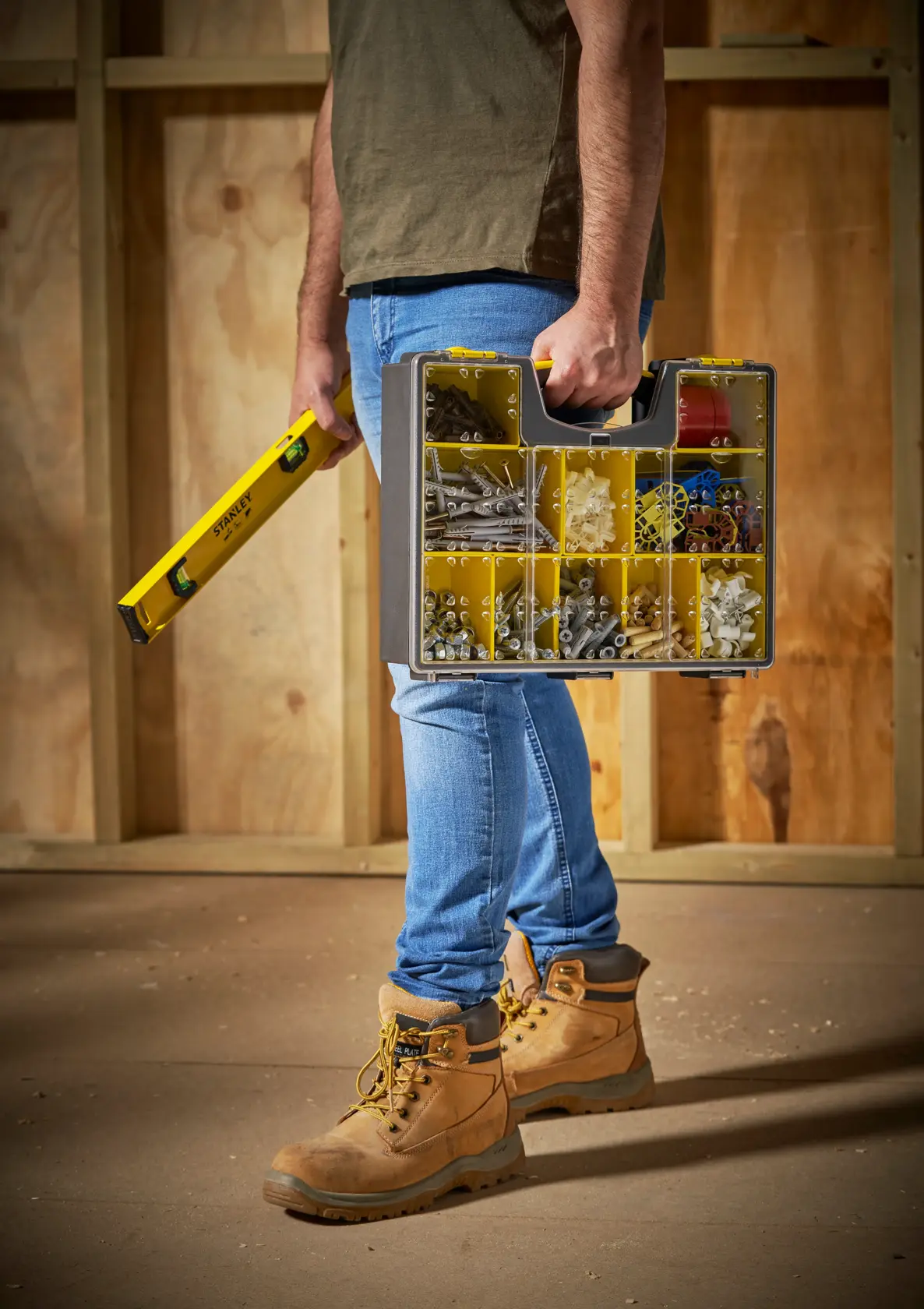 Caja organizadora STANLEY STST14710 con tapa transparente y compartimentos amarillos, llevada por persona con botas de trabajo, vista lateral.
