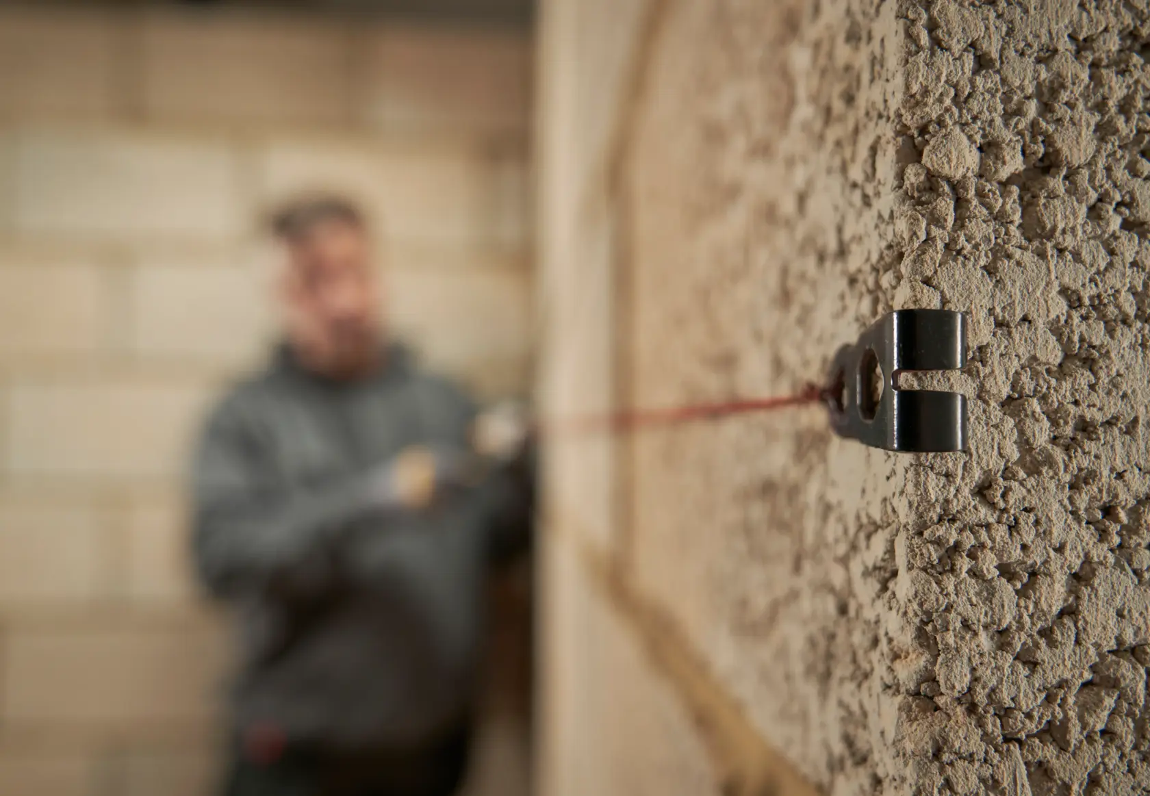 Vista de primer plano de una pared de mampostería con un bloque de línea STANLEY fijado y una línea roja tensa a lo largo de la pared. Al fondo, una persona con sudadera gris está desenfocada, preparando la línea para la alineación de la construcción.